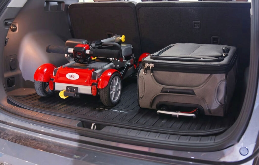 Red EV Rider Gypsy Q2 lightweight mobility scooter folded completely flat inside a car trunk next to a suitcase for easy travel storage.