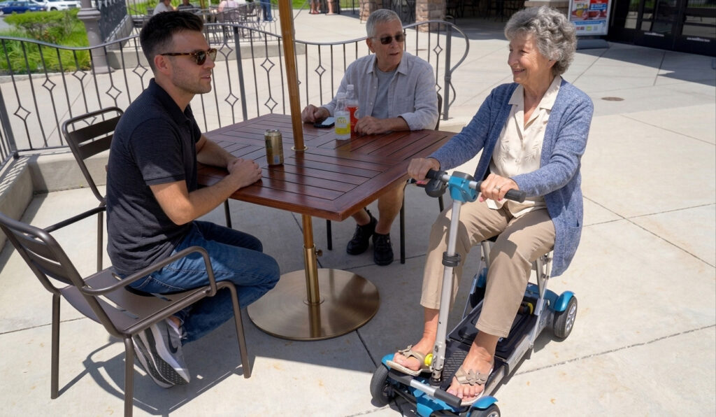 An older woman sitting on her blue EV Rider Gypsy Q2 ultra-portable scooter while enjoying drinks at an outdoor patio table with family.